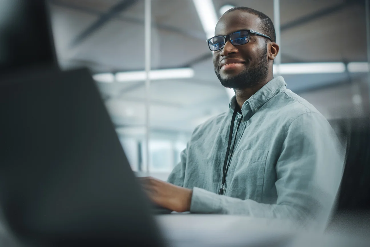 A smiling man wearing glasses and a light blue button-down shirt working at a computer in a modern office.