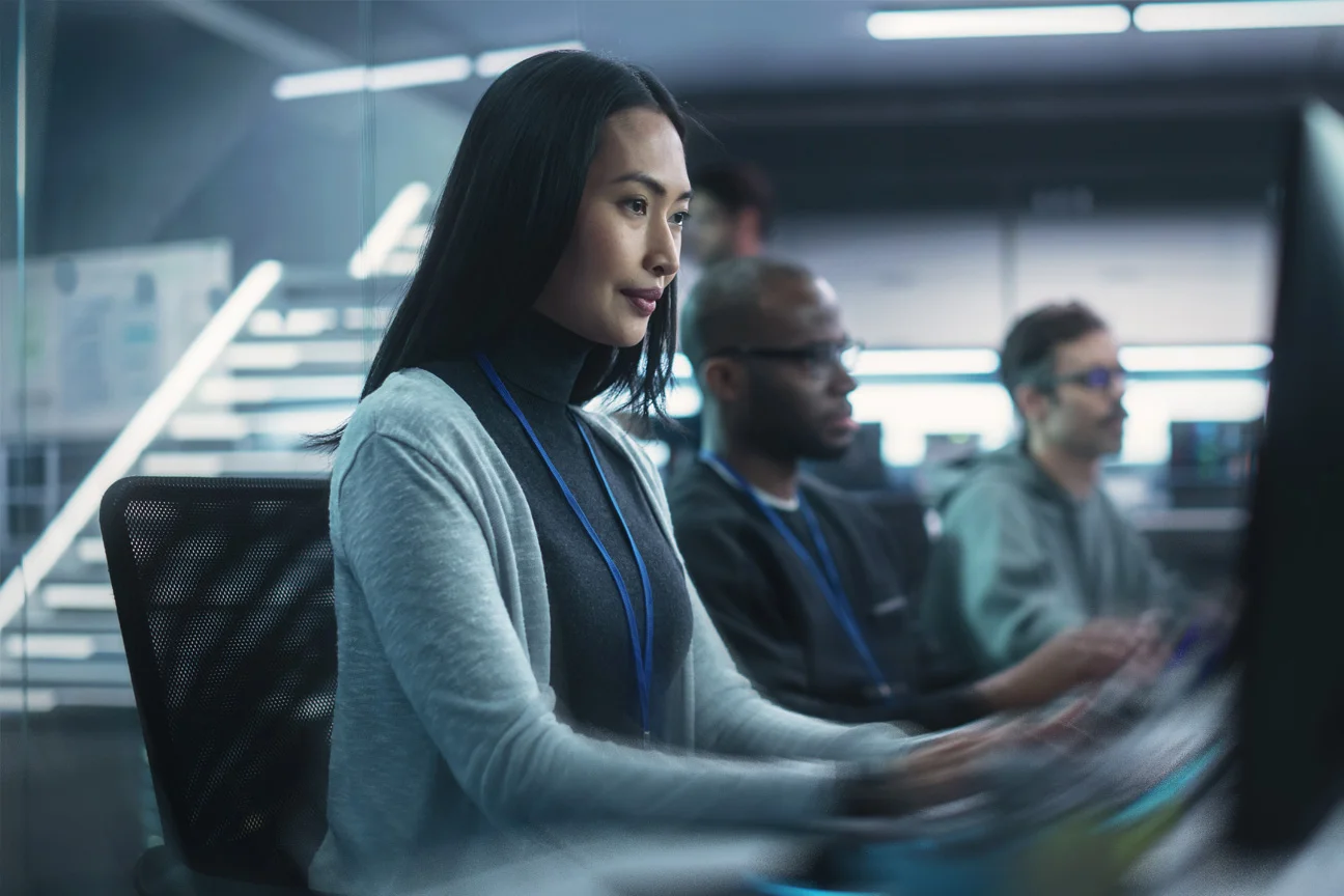 A woman with long dark hair sits focused at a computer monitor in a modern office, with colleagues working alongside her in the background.